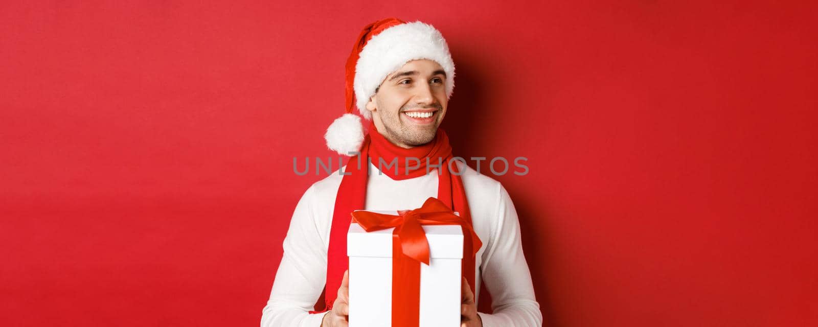 Concept of winter holidays, christmas and lifestyle. Close-up of attractive man in santa hat and scarf, holding new year gift, looking right and smiling, standing over red background.