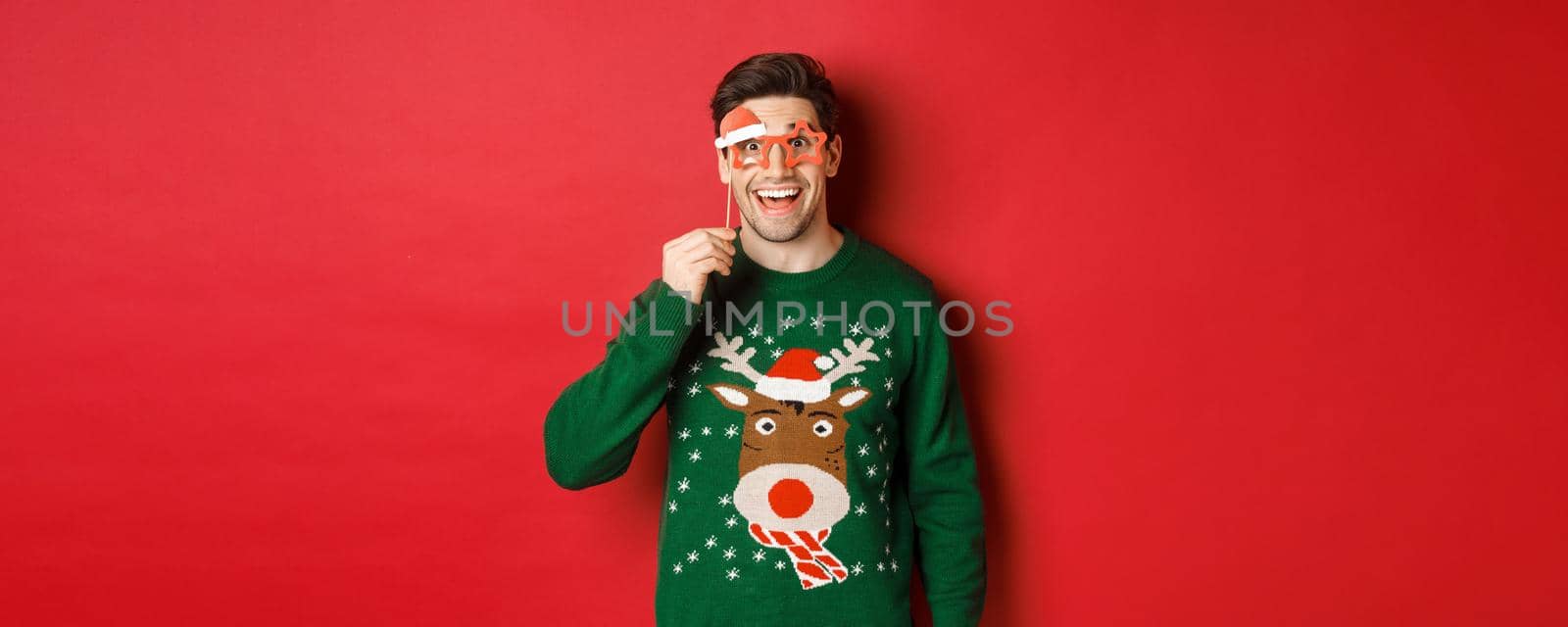 Portrait of handsome smiling man in christmas sweater and party glasses, looking surprised and happy, celebrating new year and having fun, standing against red background by Benzoix