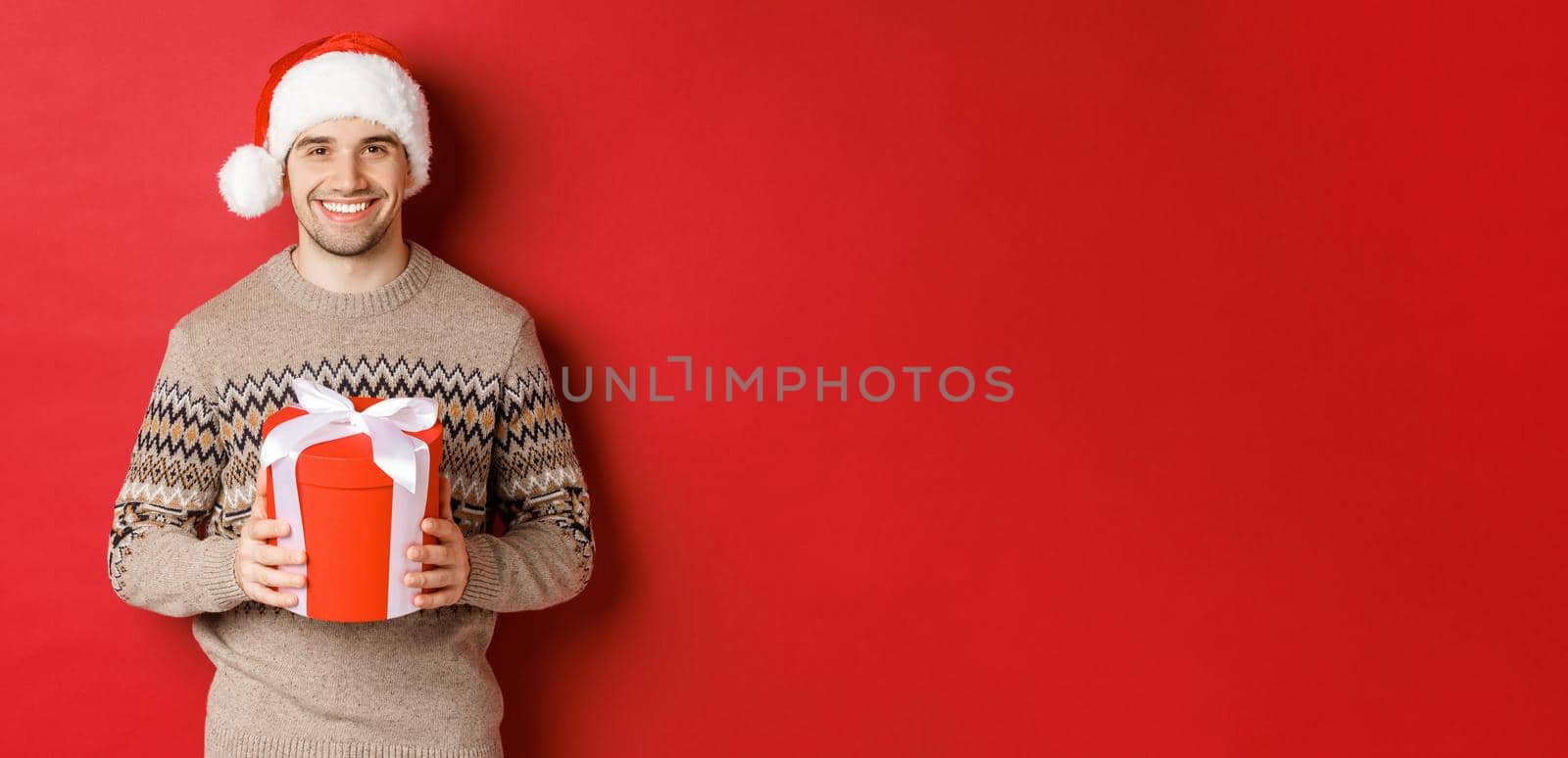 Portrait of handsome man holding a present, wishing happy holiday, standing in santa hat and christmas sweater against red background.