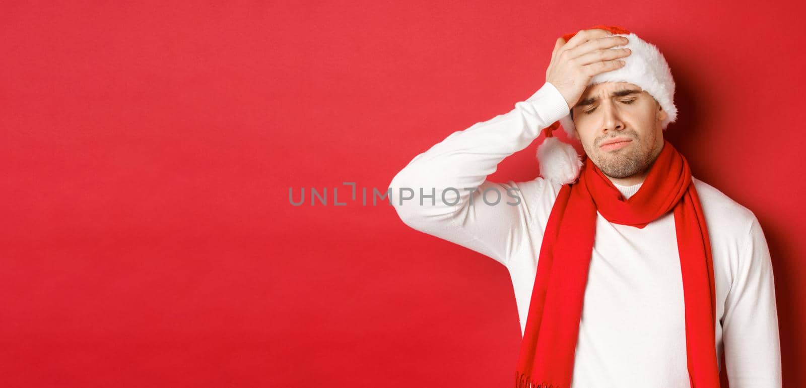 Concept of christmas, winter holidays and celebration. Portrait of sad and frustrated man in santa hat, feeling bad, standing over red background.