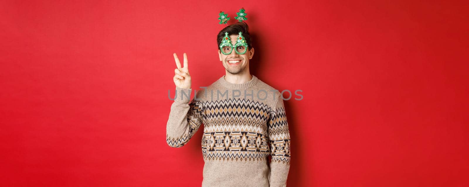 Portrait of funny and cute man in party glasses and christmas sweater, smiling and showing peace sign while posing over red background for new year advertisement.