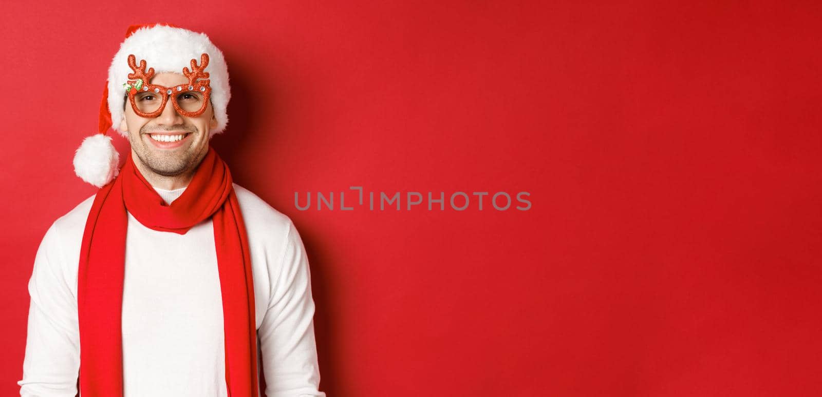 Concept of christmas, winter holidays and celebration. Cheerful handsome guy in new year party glasses and smiling, standing over red background by Benzoix