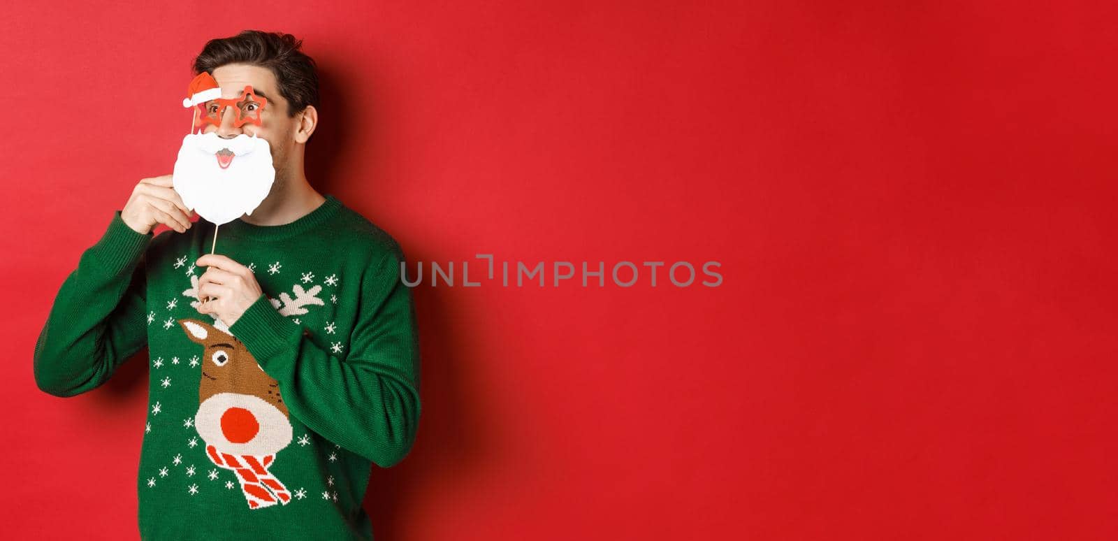 Portrait of surprised man in green christmas sweater, holding funny santa claus mask, celebrating new year, looking left amazed, sanding over red background by Benzoix