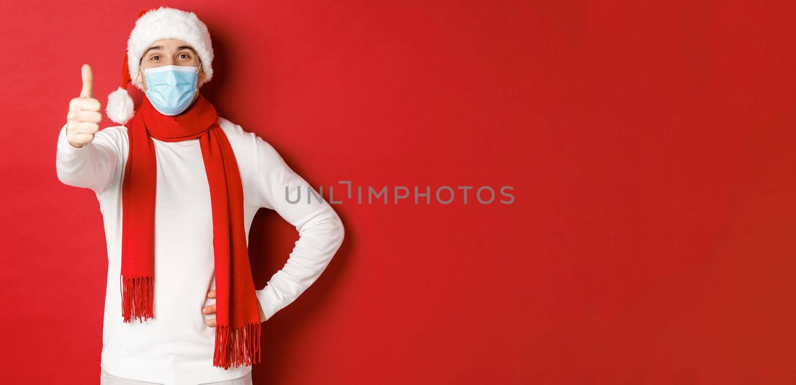 Concept of covid-19, christmas and holidays during pandemic. Cheerful handsome man in medical mask and santa hat, showing thumb-up, celebrating new year and social distancing by Benzoix