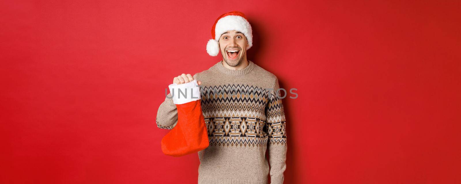 Concept of winter holidays, new year and celebration. Cheerful and surprised adult man receiving candies on Saint Nicholas day in red stocking, standing amazed over red background.