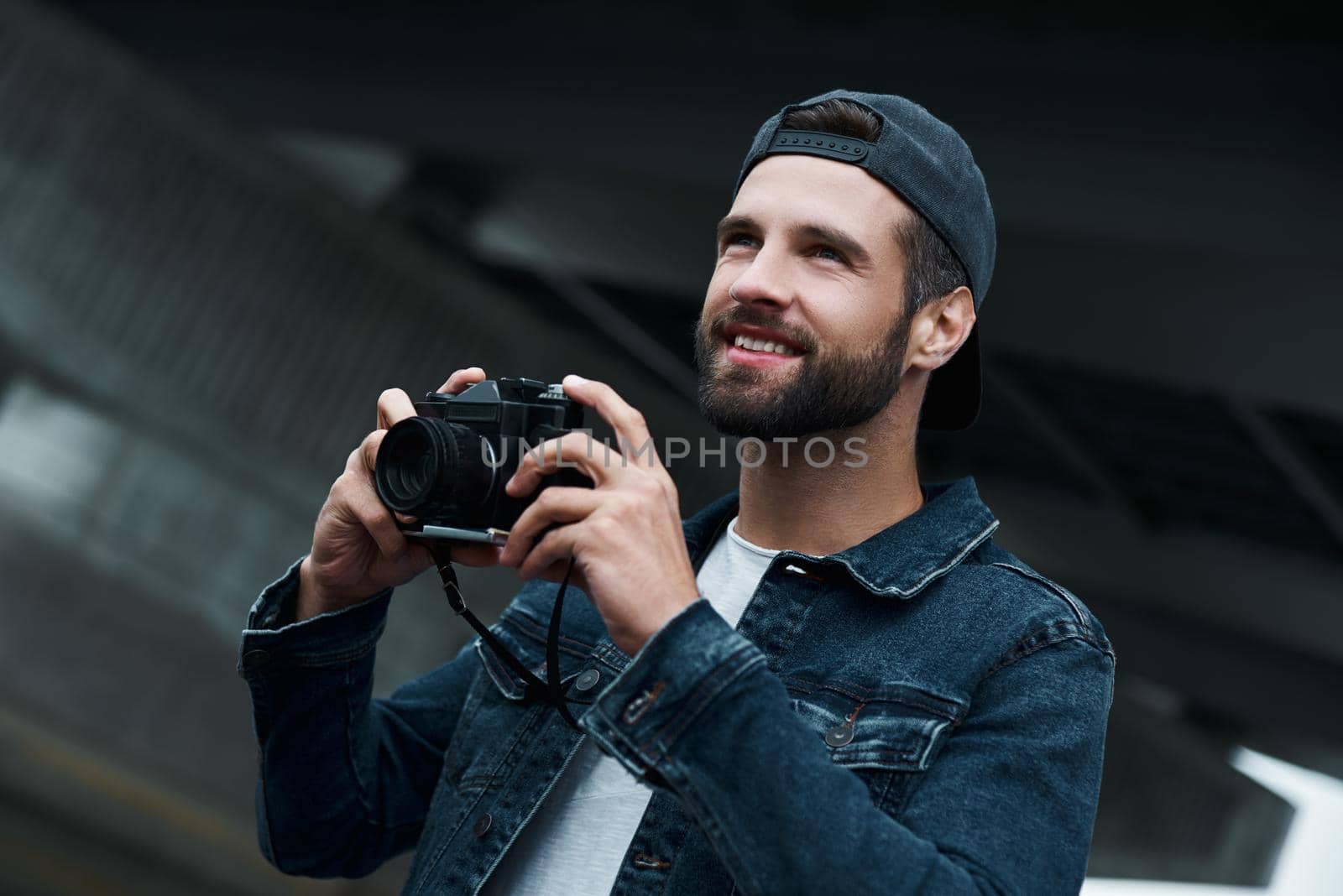 Photography hobby. Young stylish man standing on city street taking photos on camera looking forward smiling joyful close-up