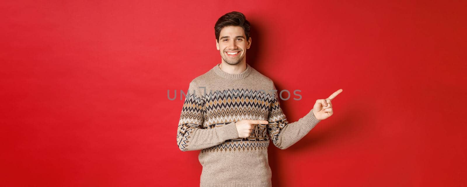 Image of attractive smiling man, wearing christmas sweater, pointing fingers right and showing new year advertisement, standing over red background.