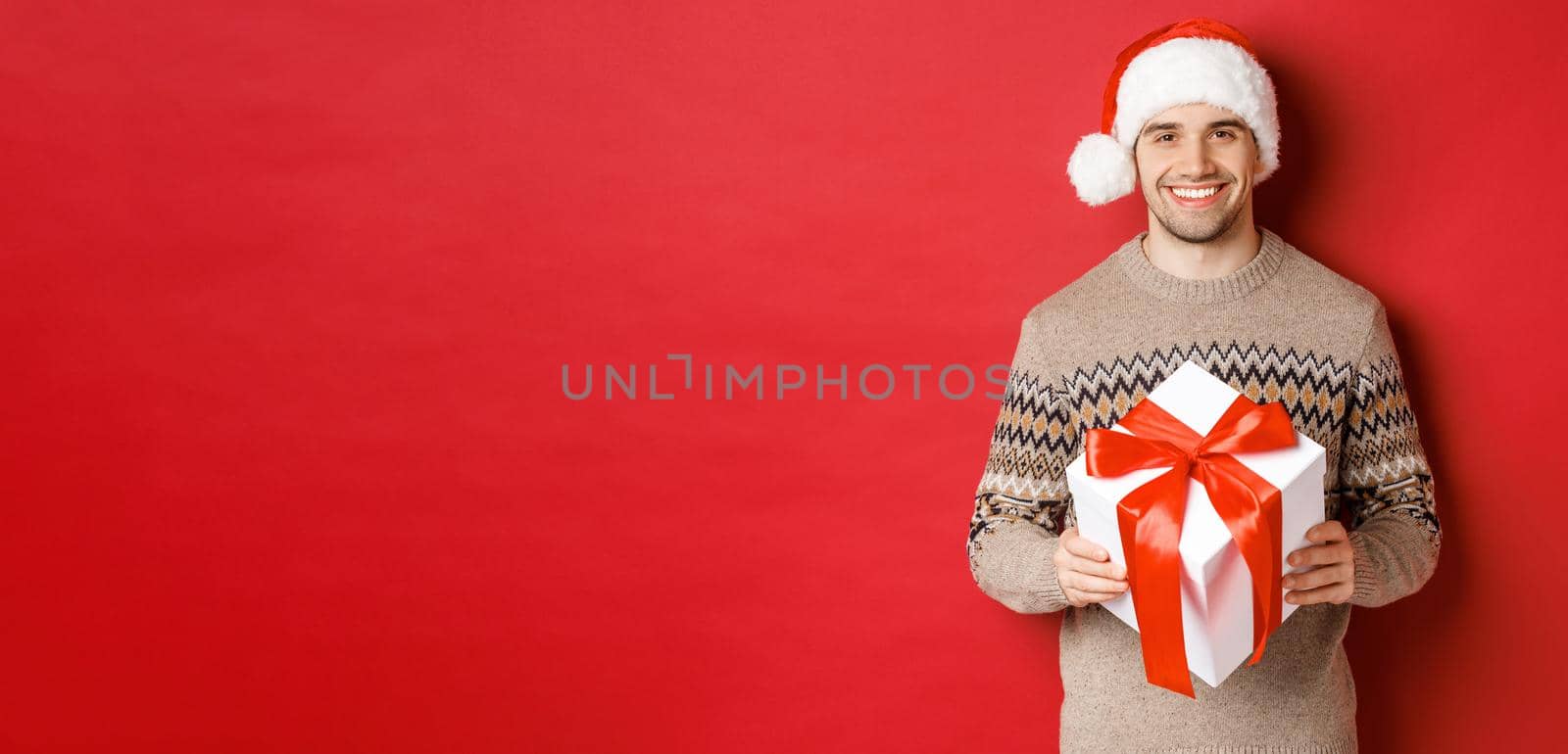 Image of handsome bearded man in winter sweater and santa hat, holding christmas gift and smiling, have a present wrapped with ribbon, standing over red background.