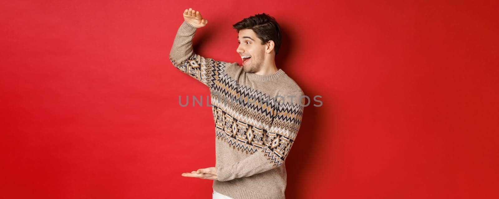 Portrait of happy young man in christmas sweater, showing large present, smiling and looking amazed at cool gift, standing over red background by Benzoix