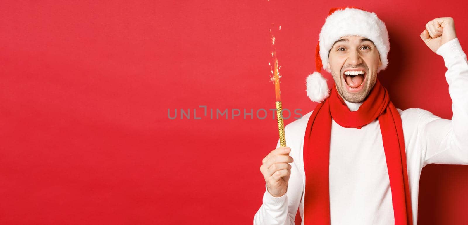 Concept of christmas, winter holidays and celebration. Handsome man celebrating new year and having fun, holding sparkler and smiling, wearing santa hat, standing over red background by Benzoix