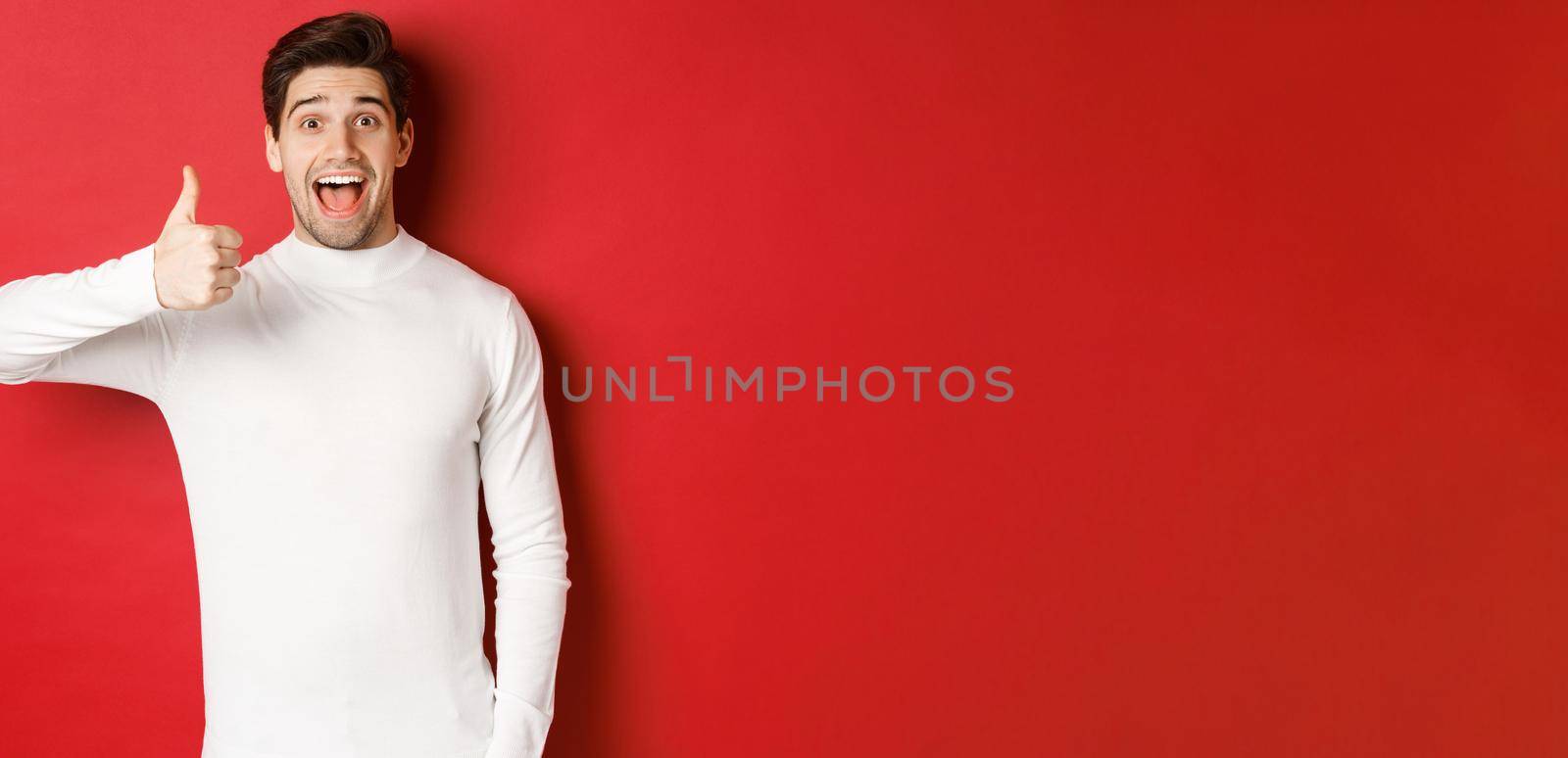 Concept of winter holidays, christmas and lifestyle. Excited good-looking man in white sweater, like good product, showing thumb-up in approval and smiling amazed, red background by Benzoix