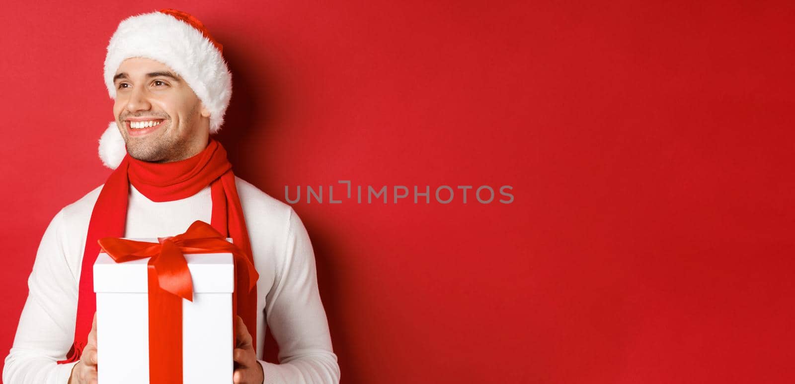 Concept of winter holidays, christmas and lifestyle. Close-up of attractive guy in santa hat and scarf, smiling and looking left while holding new year present, standing over red background.