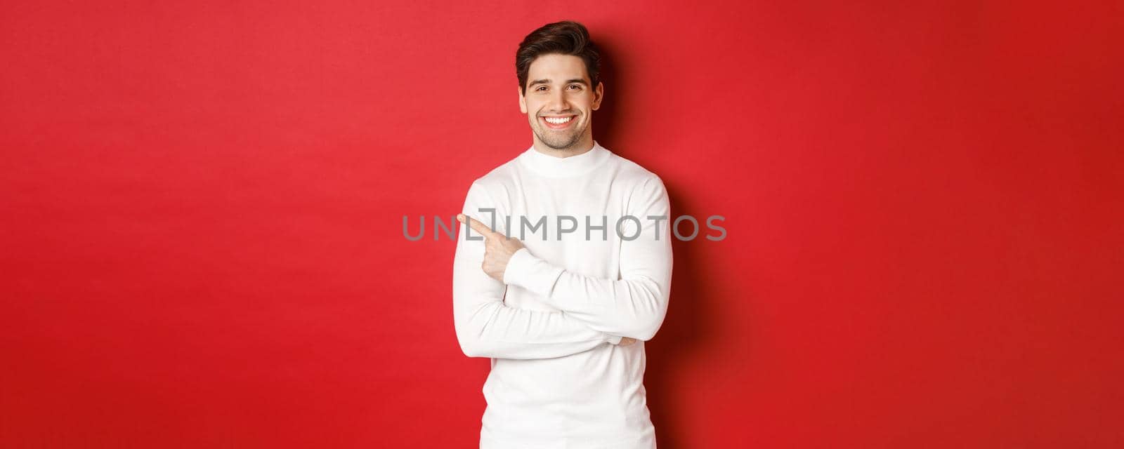 Concept of winter holidays, christmas and lifestyle. Portrait of attractive man in white sweater, pointing finger at upper left corner and smiling, showing logo on red background.