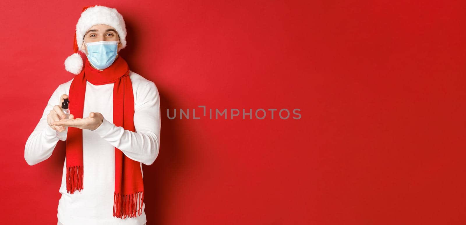 Concept of covid-19, christmas and holidays during pandemic. Happy young man in santa hat and medical mask, sanitize hands with antiseptic and smiling, standing over red background.