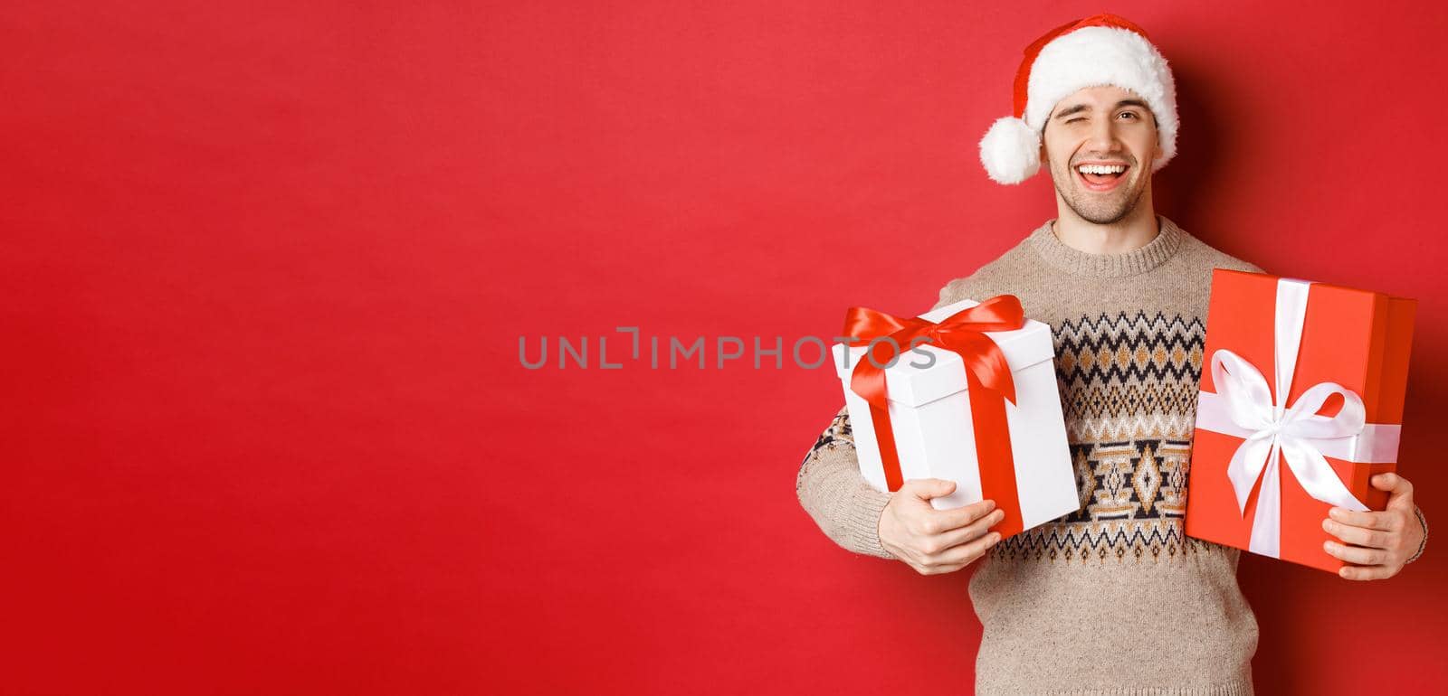 Concept of winter holidays, new year and celebration. Portrait of confident and cheeky young man prepared gifts for christmas, winking and holding presents, standing over red background by Benzoix