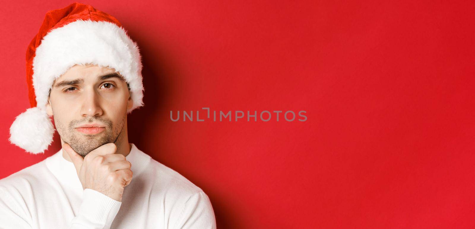 Close-up of thoughtful handsome man in santa hat, frowning and looking at camera, thinking about something, standing over red background.