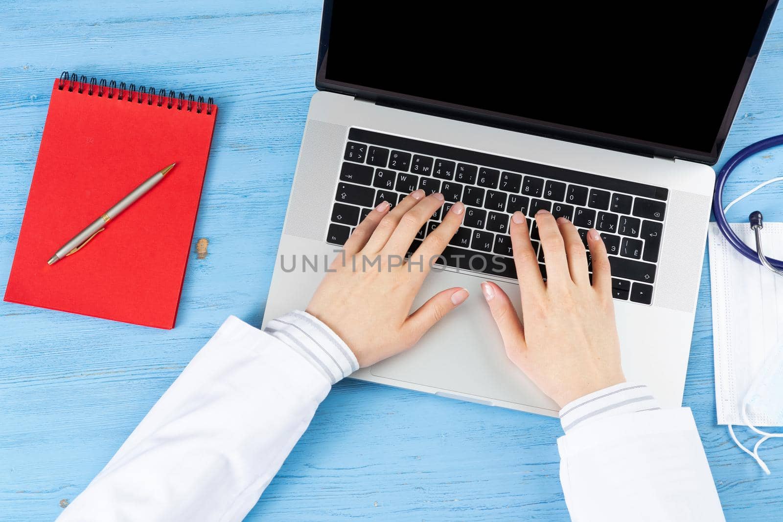 Top view of doctor hands typing at laptop computer. Therapist in white coat sitting at blue wooden desk. Examination and consultation in clinic. Medical application and online healthcare services.