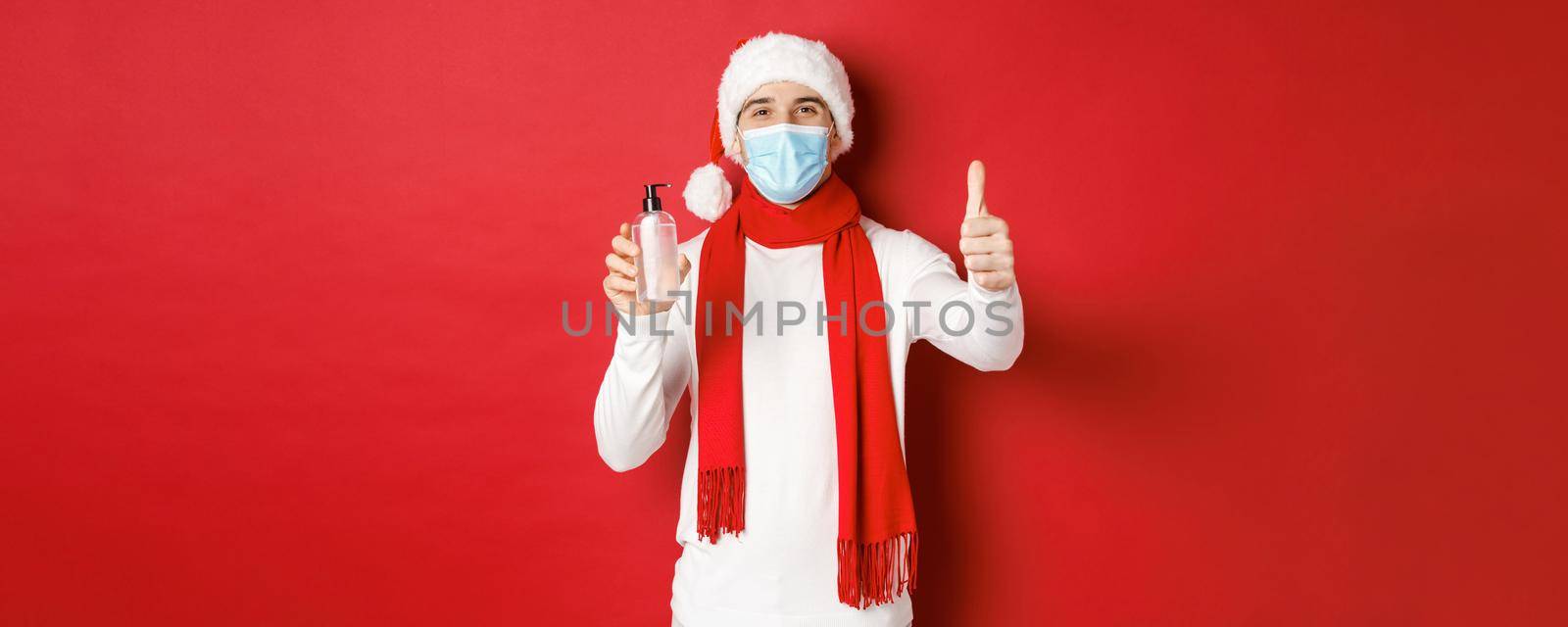 Concept of covid-19, christmas and holidays during pandemic. Happy and satisfied man in santa hat and medical mask, showing thumb-up in approval and recommending hand sanitizer by Benzoix