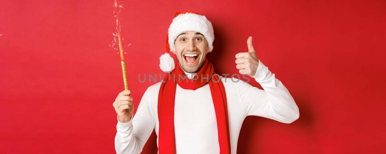 Concept of christmas, winter holidays and celebration. Handsome man celebrating new year and having fun, holding sparkler and showing thumb-up, standing over red background.