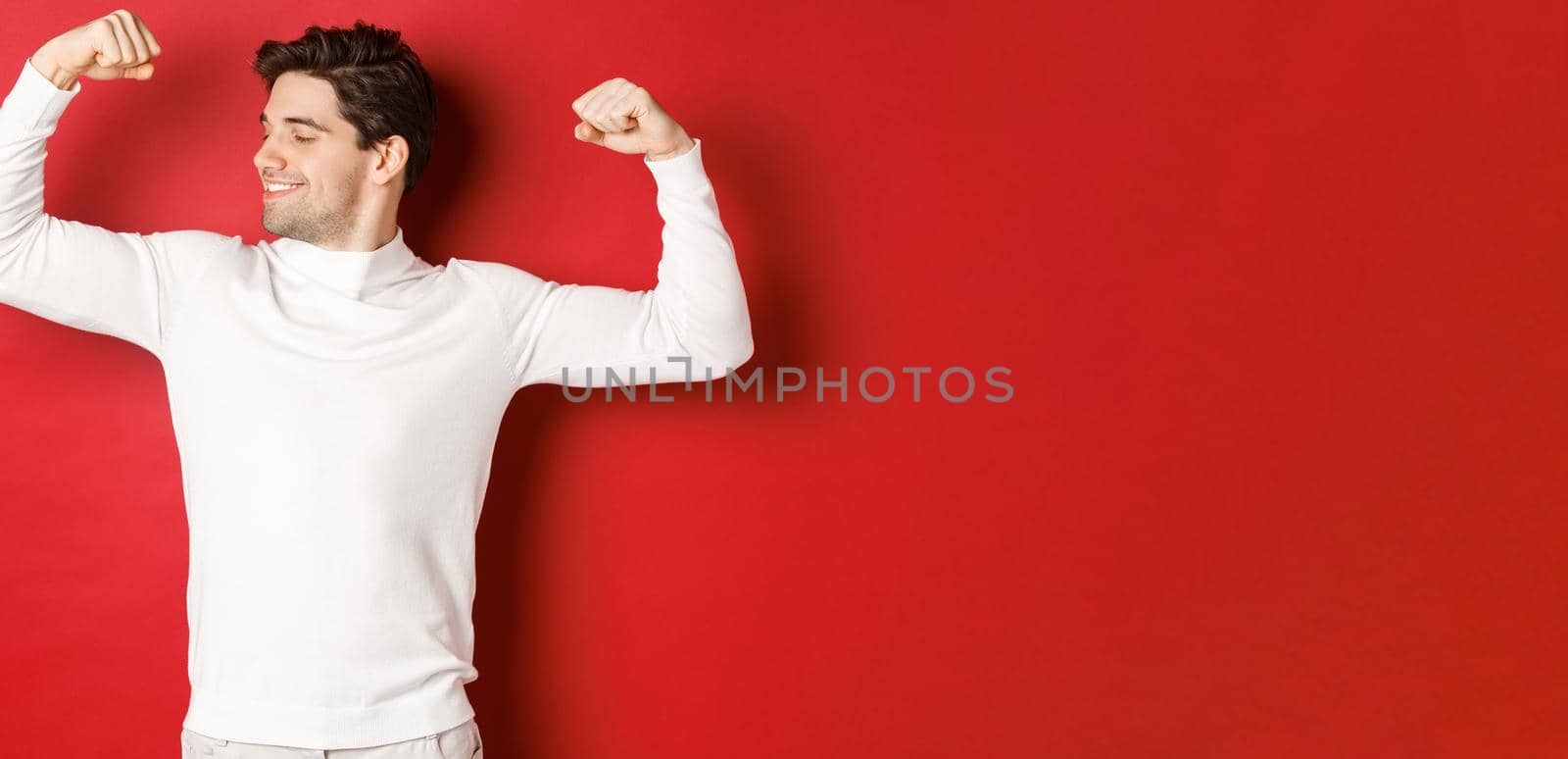 Portrait of smiling handsome man in white sweater, flexing biceps and bragging with strength, show-off strong muscles after workout, standing over red background by Benzoix