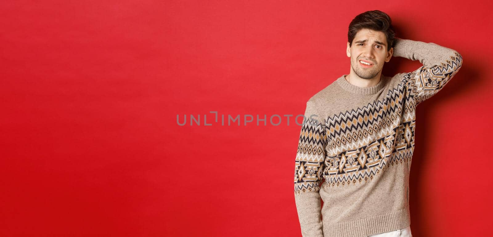 Portrait of confused man in christmas sweater, scratching back of head and looking clueless about new year presents, standing over red background.