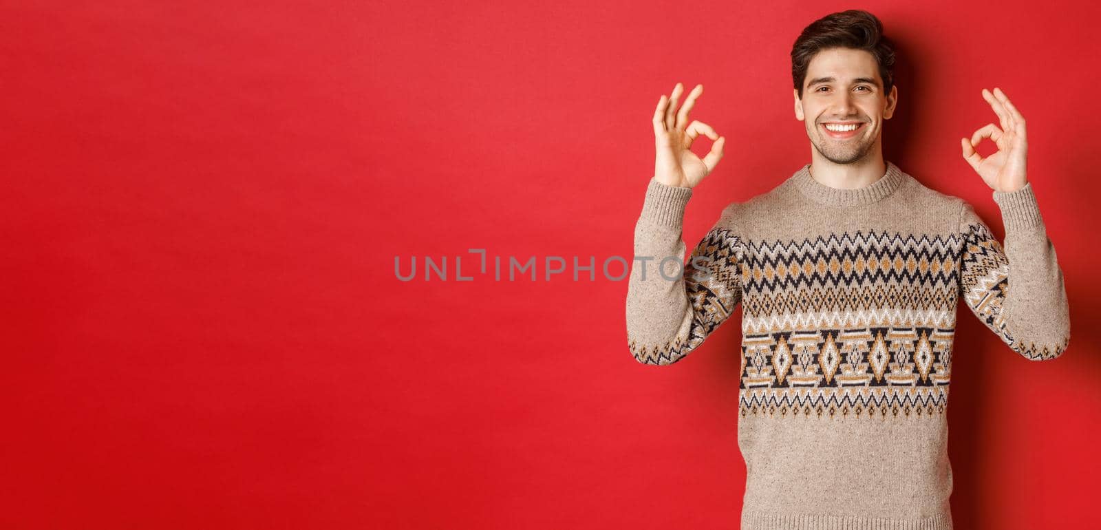 Concept of christmas celebration, winter holidays and lifestyle. Satisfied handsome man in sweater, showing okay signs and smiling pleased, recommending something, red background.