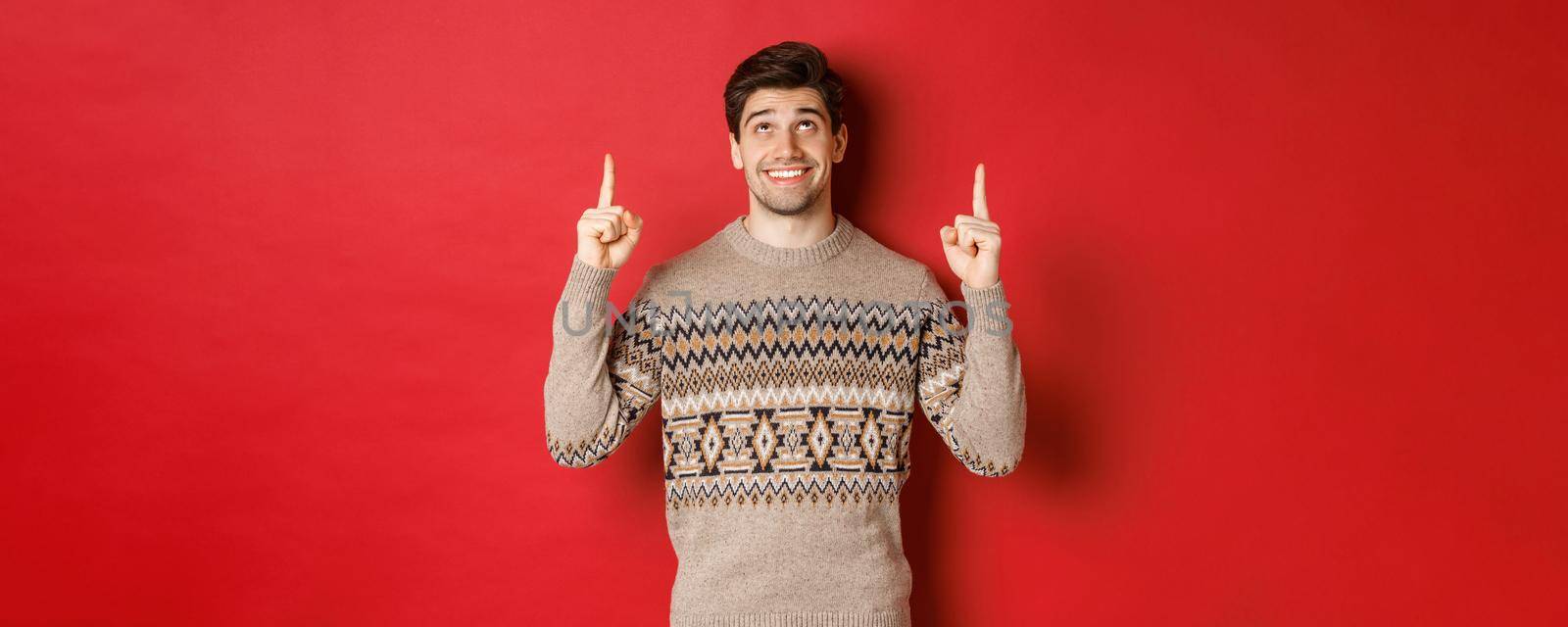 Portrait of dreamy handsome guy thinking about christmas holidays, wearing winter sweater, pointing and looking up at new year promo offer, standing over red background by Benzoix