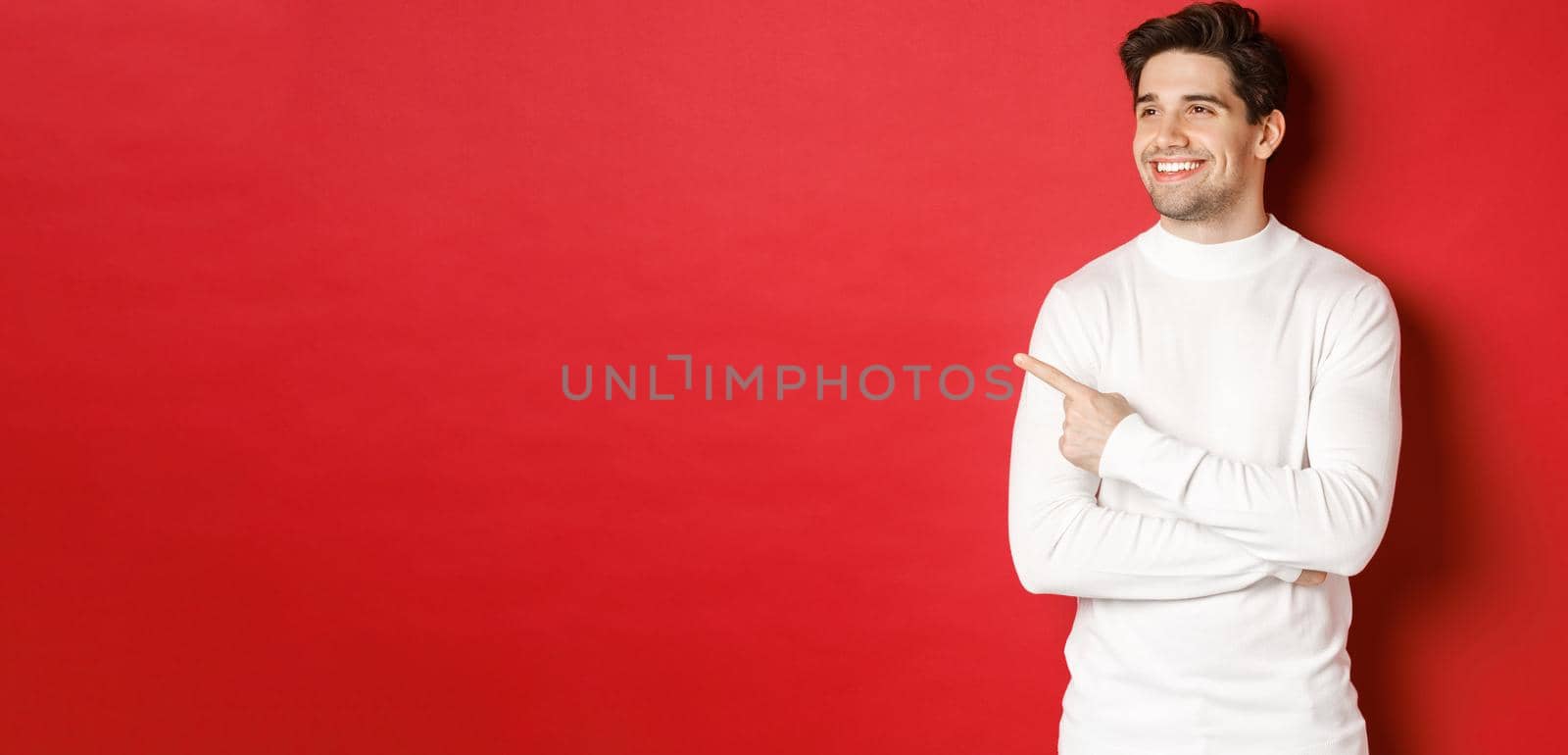Concept of winter holidays, christmas and lifestyle. Handsome adult man in white sweater, looking and pointing left with pleased smile, showing new year advertisement, red background by Benzoix
