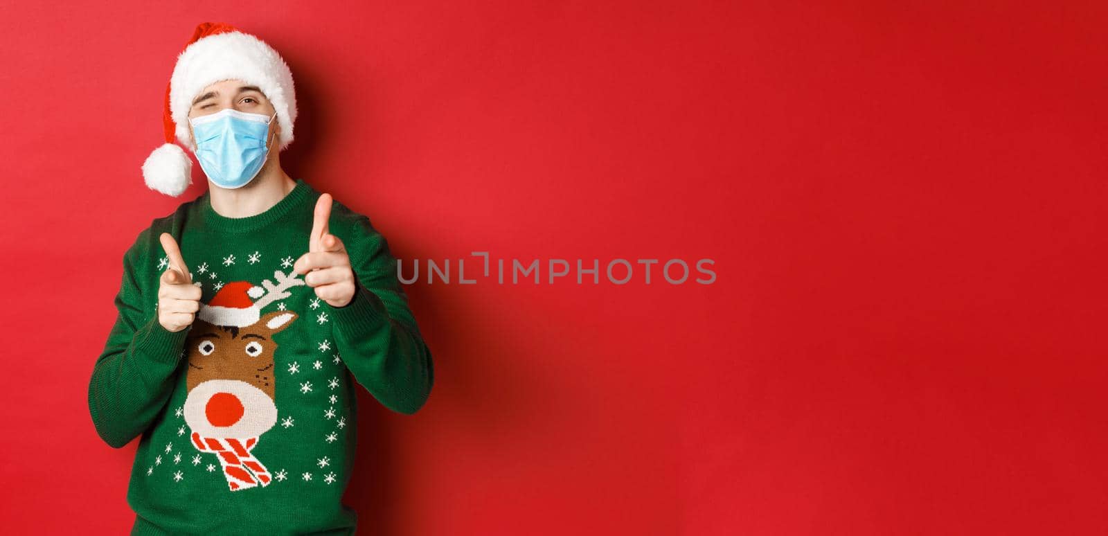Concept of new year, covid-19 and social distancing. Cheerful man in christmas sweater, medical mask and santa hat, pointing fingers at camera, wishing happy holidays, standing over red background.