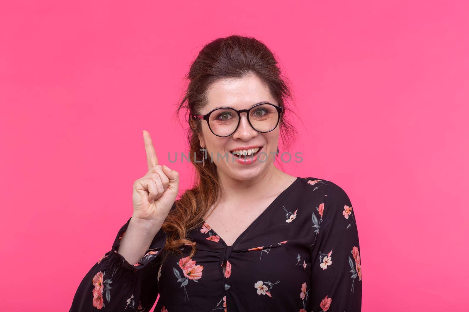 Portrait of a young beautiful romantic young female student with glasses showing her finger up posing on a pink background. Space for advertising. Concept of new ideas