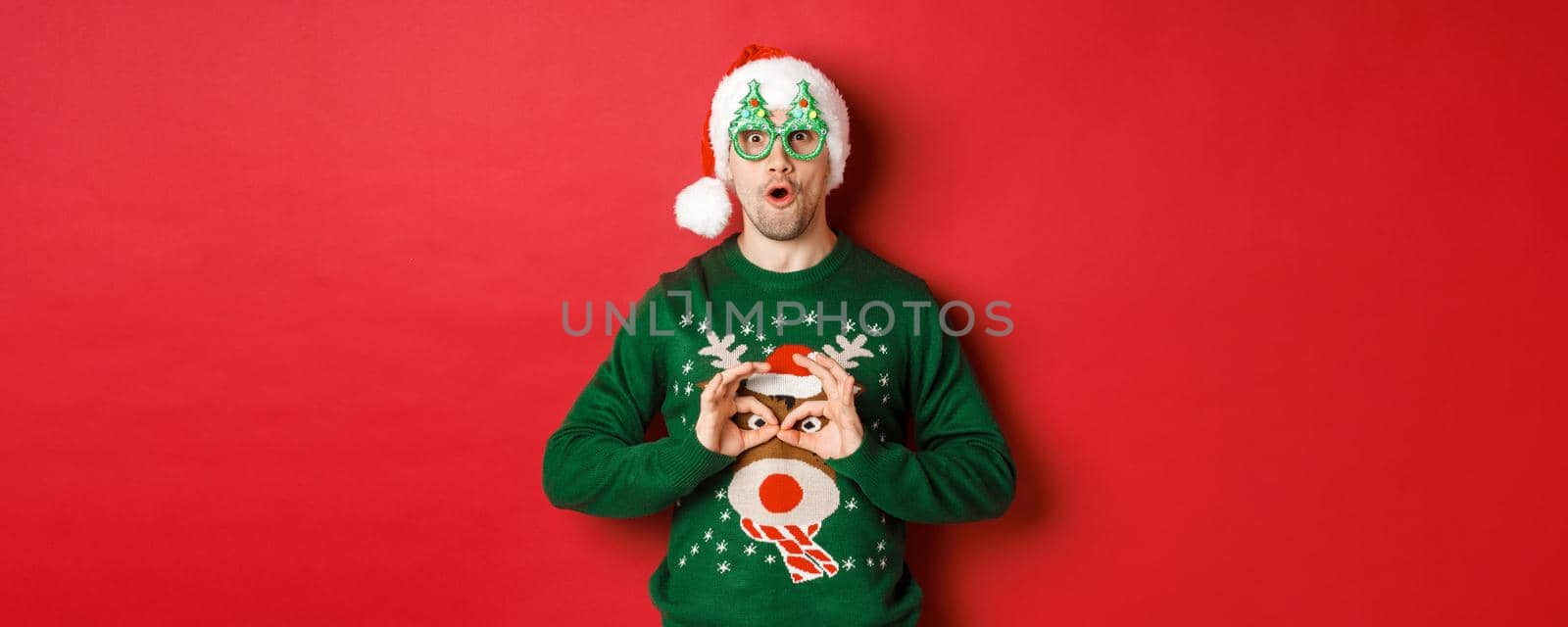 Portrait of carefree handsome man in santa hat and party glasses, making fun of his christmas sweater, looking happy over red background.