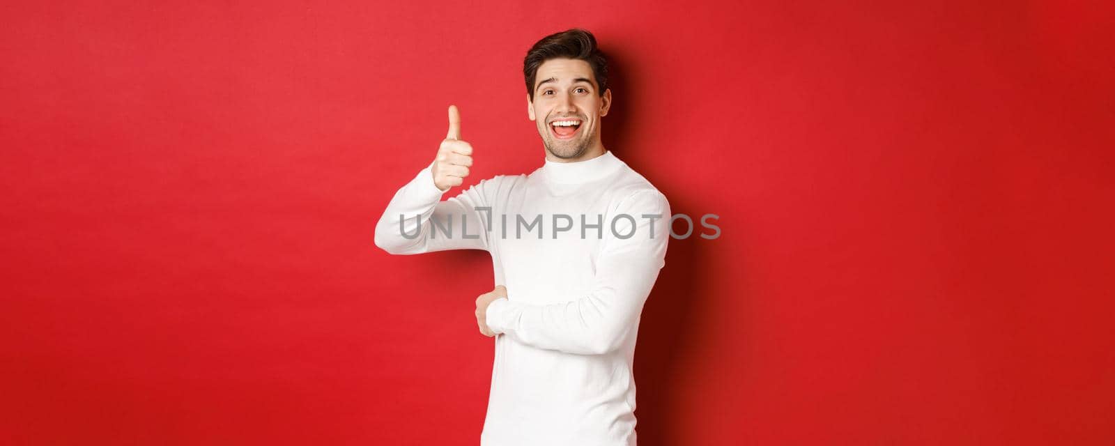 Concept of winter holidays, christmas and lifestyle. Portrait of cheerful, attractive man in white sweater, showing thumbs-up in approval, like good idea, standing over red background.