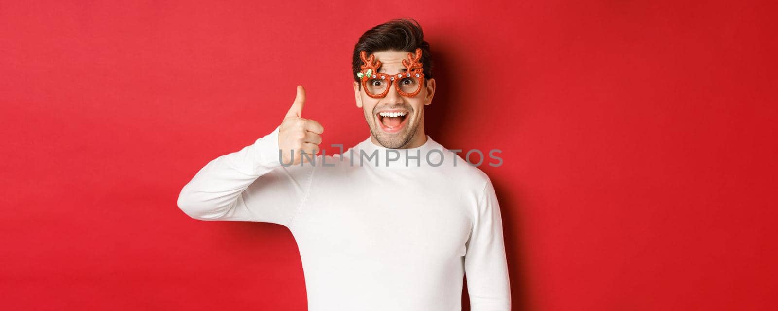 Close-up of handsome young man in party glasses and white sweater, showing thumb-up in approval, enjoying new year holidays, standing over red background by Benzoix