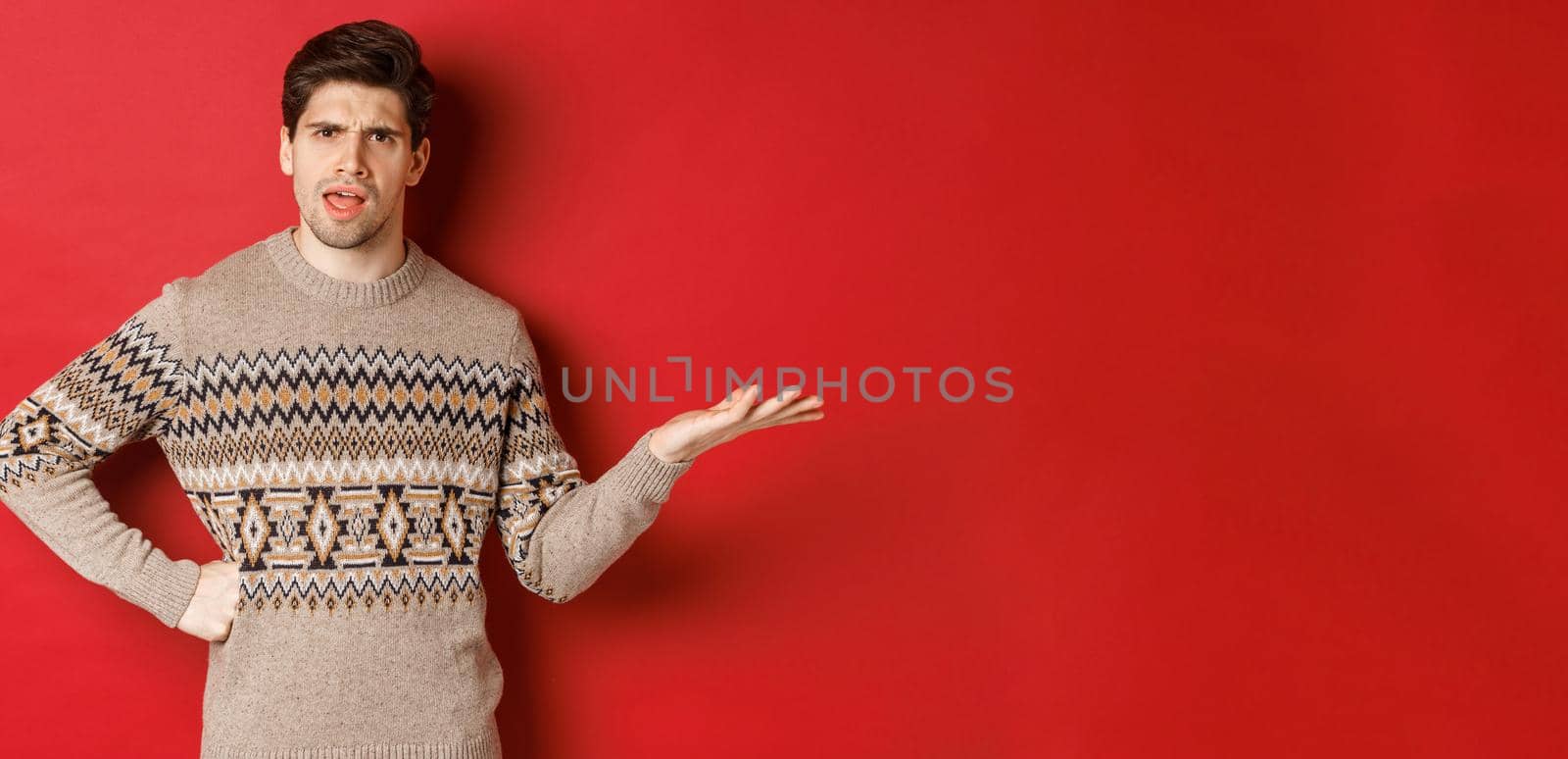 Image of disappointed and confused handsome man, wearing christmas sweater, raising hand and frowning, asking you to explain something, standing over red background.