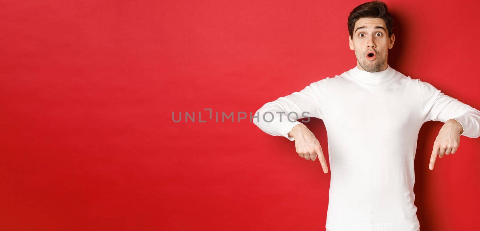 Image of surprised attractive guy in white sweater, showing announcement, pointing fingers down and looking amazed, standing over red background.