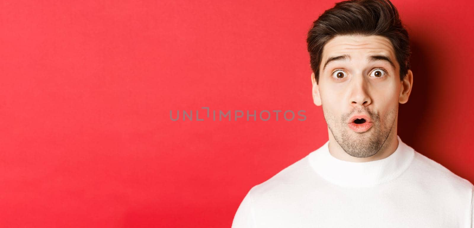Close-up of surprised handsome man in white sweater, open mouth and looking wondered, hear amazing news, standing over red background by Benzoix