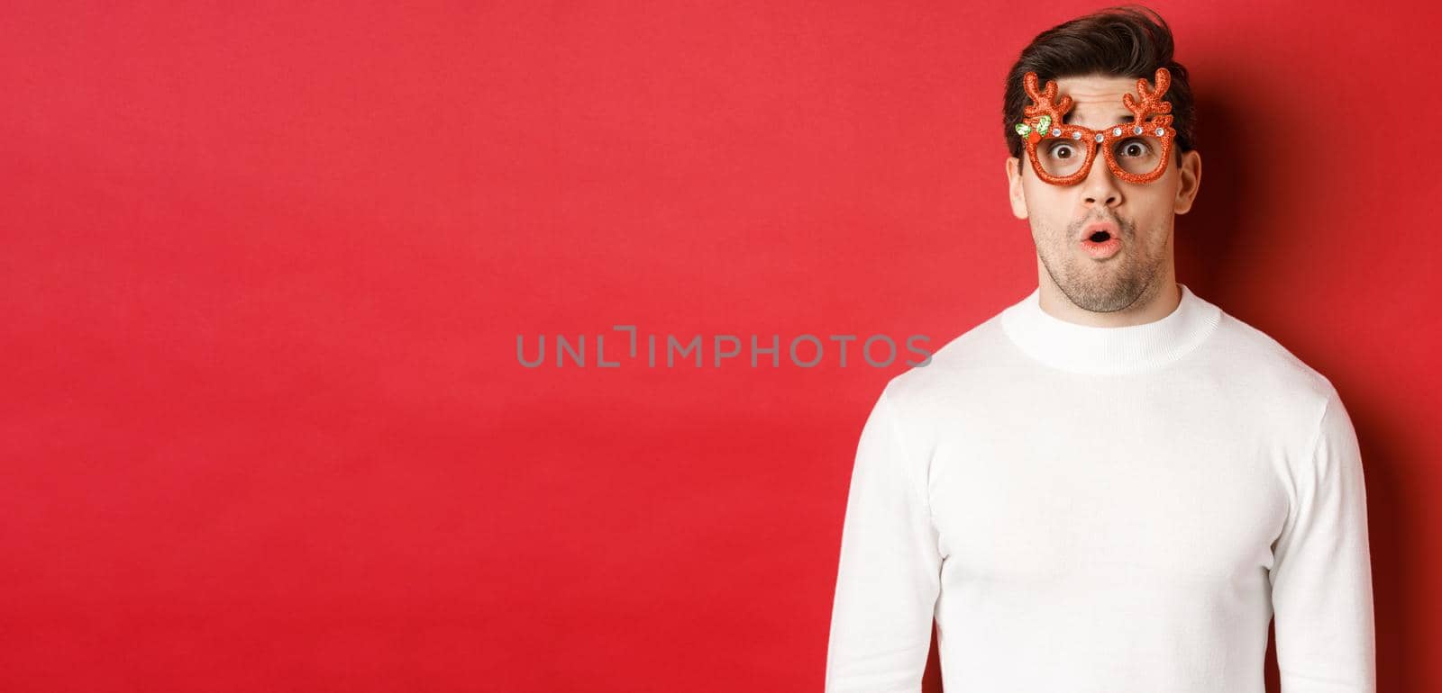 Close-up of surprised and impressed man in white sweater, party glasses, saying wow and looking amazed at camera, standing over red background.