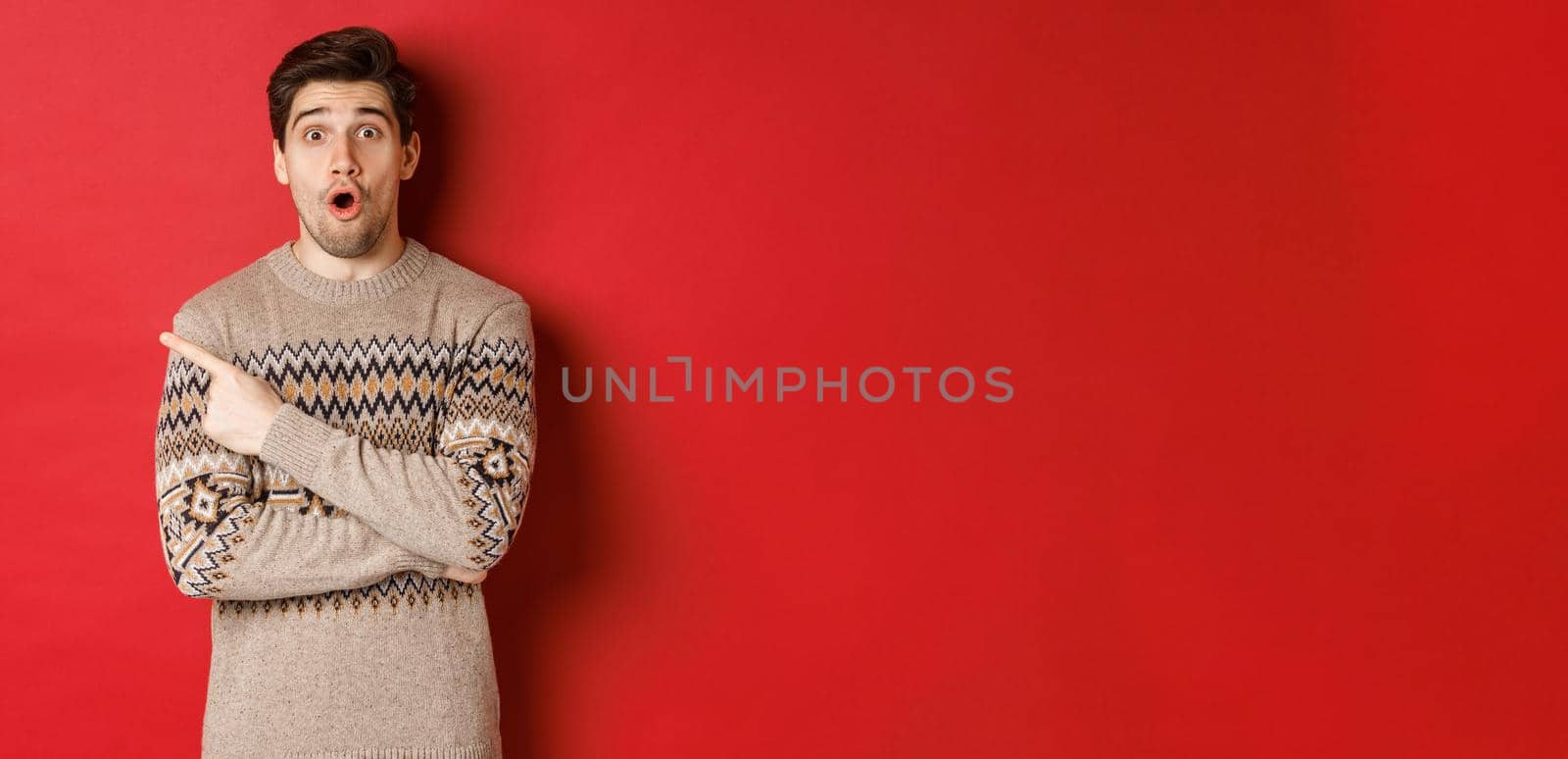 Image of impressed handsome guy, showing awesome christmas promo offer, pointing finger at upper left corner and looking amazed, standing in xmas sweater over red background.