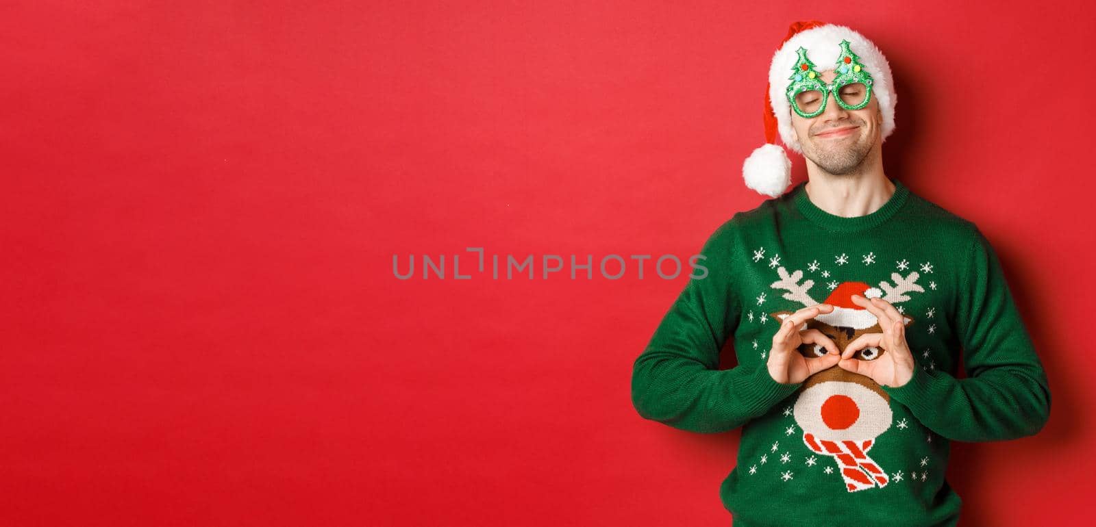 Image of happy smiling man in party glasses and santa hat, fooling around with funny christmas sweater, celebrating winter holidays, standing over red background by Benzoix