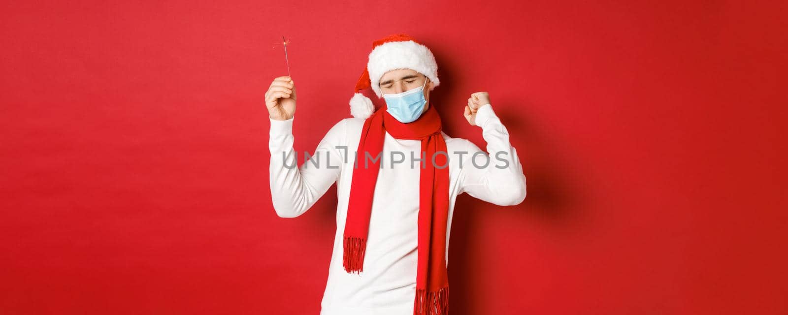Concept of covid-19, christmas and holidays during pandemic. Happy man celebrating new year at party, wearing medical mask and santa hat, dancing with sparkler against red background by Benzoix