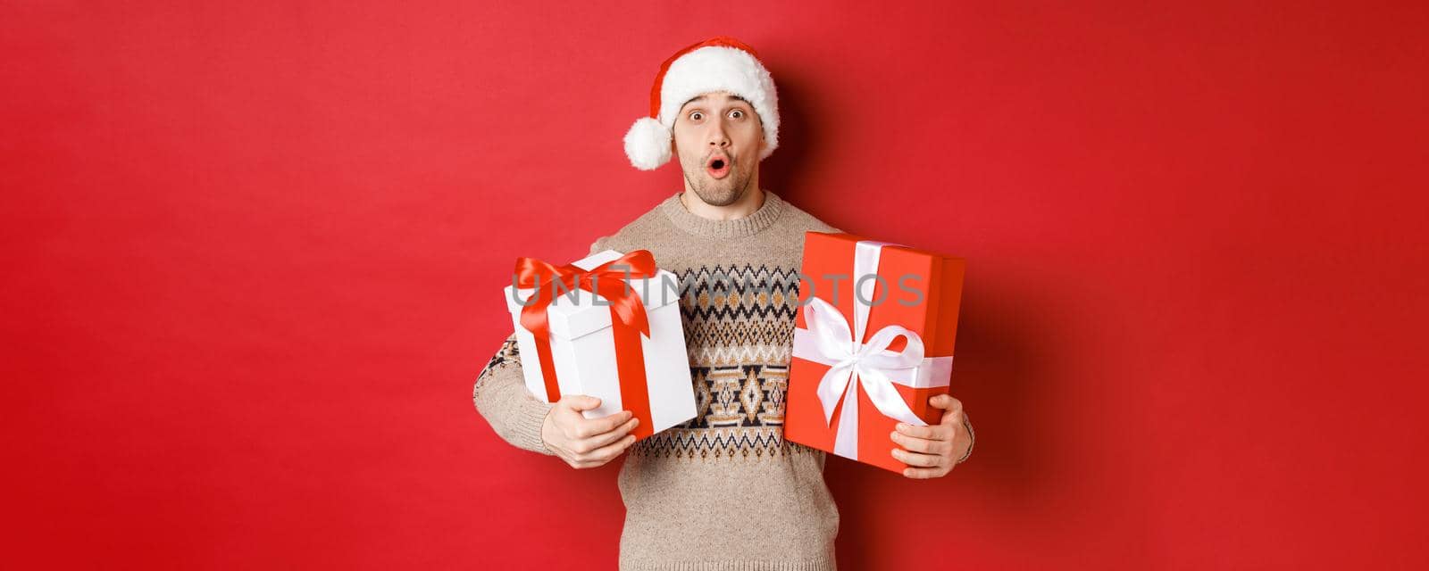 Concept of winter holidays, new year and celebration. Image of surprised attractive guy in santa hat and christmas sweater, receiving gifts, holding presents and looking amazed.