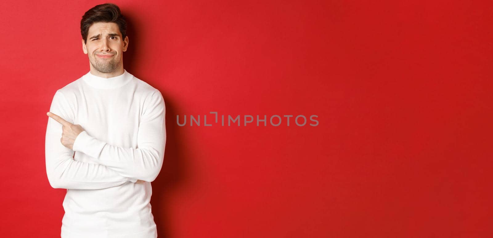 Concept of winter holidays, christmas and lifestyle. Image of skeptical young man in white sweater, dont like something, pointing finger left and looking with dismay, standing over red background.
