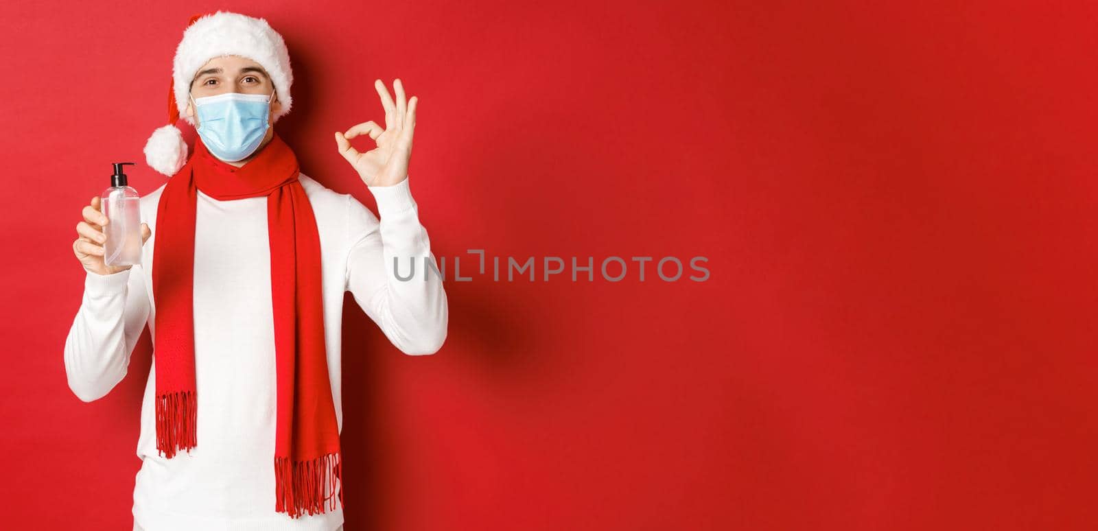Concept of covid-19, christmas and holidays during pandemic. Attractive man in santa hat and medical mask, showing okay sign while recommending hand sanitizer, standing over red background by Benzoix