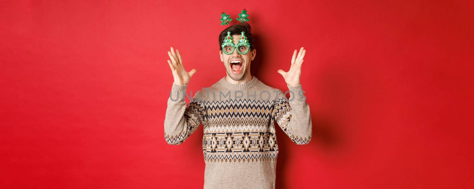 Image of excited and amazed caucasian guy in party glasses, christmas sweater, raising hands up and making big announcement, enjoying new year celebration, red background.