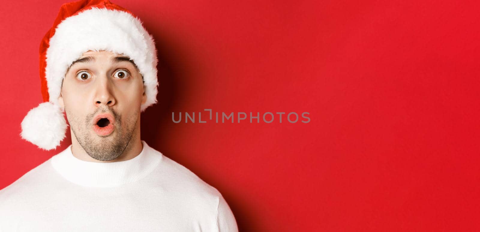 Close-up of attractive surprised man in santa hat, saying wow and looking at something interesting, standing against red background.