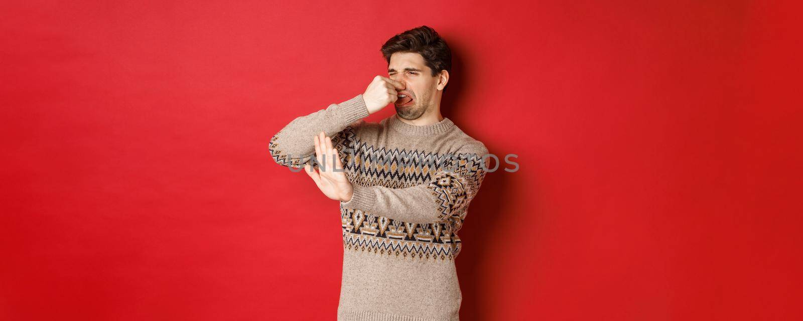 Image of disgusted handsome man in winter sweater, rejecting something with bad smell, shut nose and raising hand to decline, standing over red background.