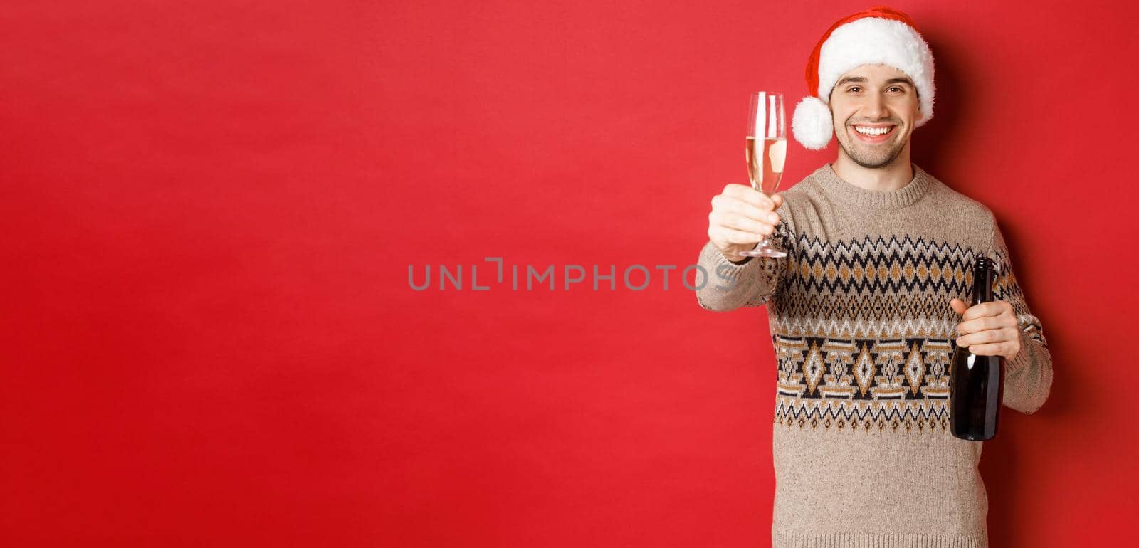 Concept of winter holidays, new year and celebration. Portrait of handsome man in santa hat and sweater, holding champagne, raising glass and saying cheers on christmas party.