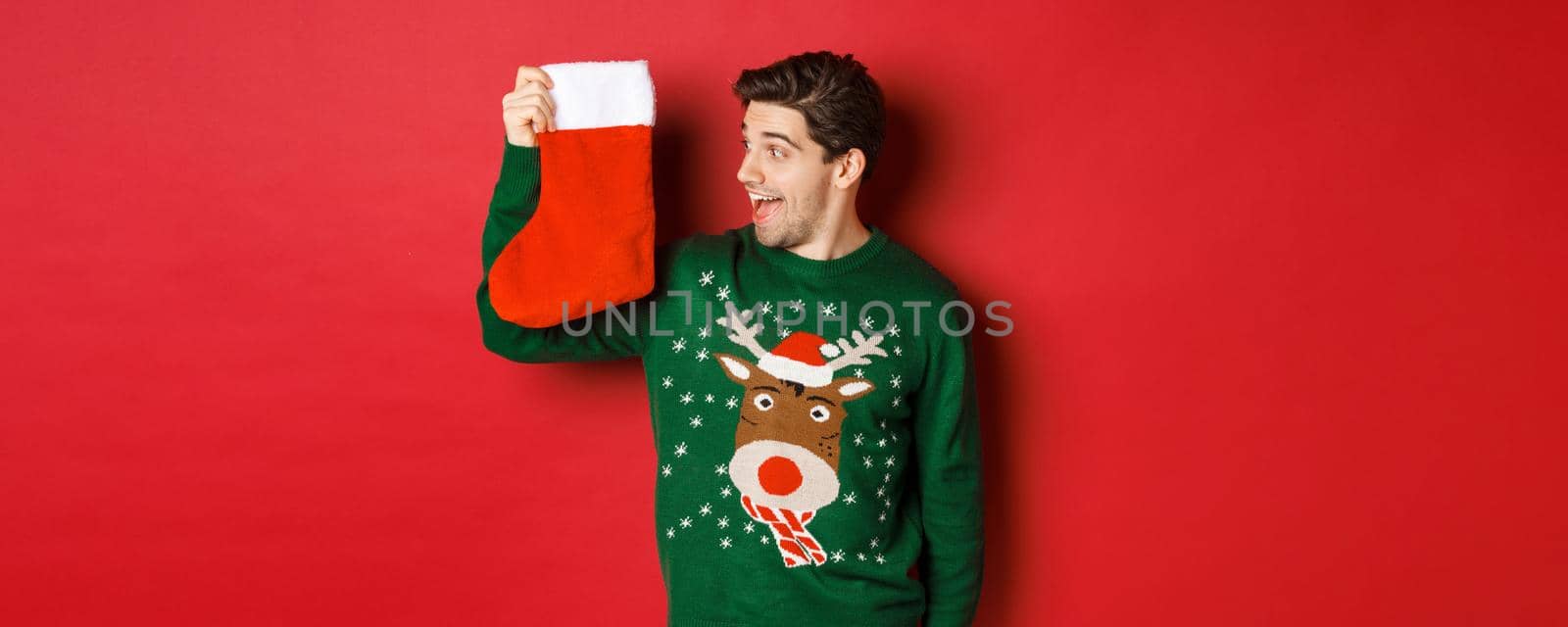 Image of surprised and amused man in green sweater, looking at christmas stocking with presents and smiling, standing over red background.