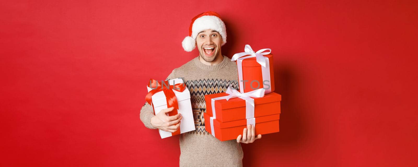 Concept of winter holidays, new year and celebration. Image of happy guy enjoying christmas, holding pile of presents and smiling amused, standing over red background by Benzoix