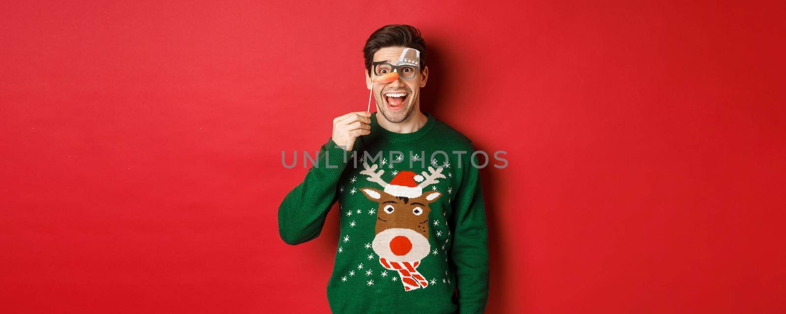 Portrait of amused handsome man in christmas sweater, holding funny party mask and smiling, celebrating winter holidays, standing over red background.